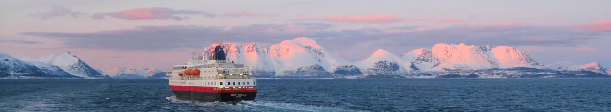 Ein Schiff von Hurtigruten Das Original fährt durch die weite norwegische Fjordlandschaft vor einer majestätischen Kulisse aus schneebedeckten Bergen, die von der tiefstehenden Sonne in ein sanftes Rosa getaucht werden.