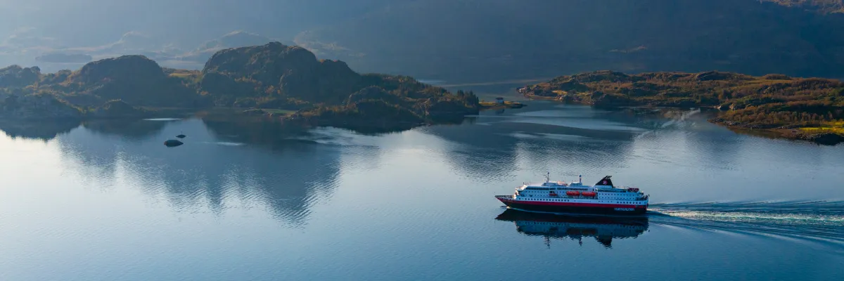 Ein klassisches Schiff von Hurtigruten Das Original gleitet durch das spiegelglatte, tiefblaue Wasser eines norwegischen Fjords, umgeben von sanften grünen Hügeln und felsigen Inseln in einer friedlichen Lichtstimmung.