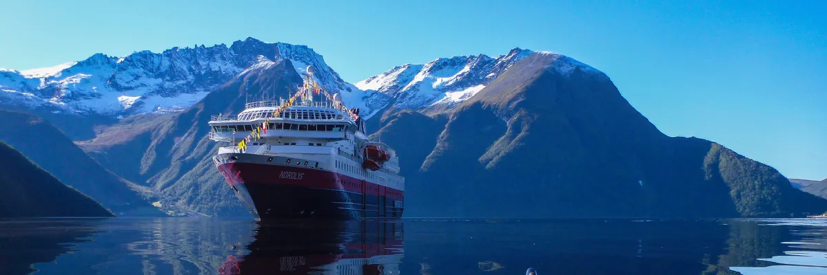 Das Postschiff MS Nordlys von Hurtigruten Das Original in einer Frontalansicht, wie es ruhig in einem norwegischen Fjord liegt. Im Hintergrund ragen gewaltige, schneebedeckte Berggipfel unter einem strahlend blauen Himmel auf.