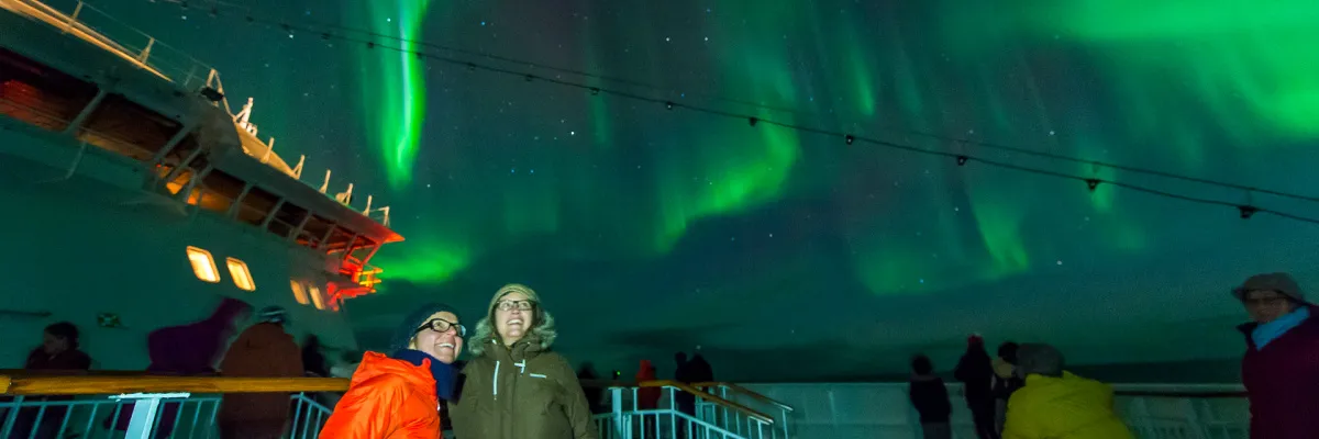 Gäste an Bord eines Hurtigruten Schiffes beobachten die magischen grünen Nordlichter am Nachthimmel während einer Norwegen Seereise.