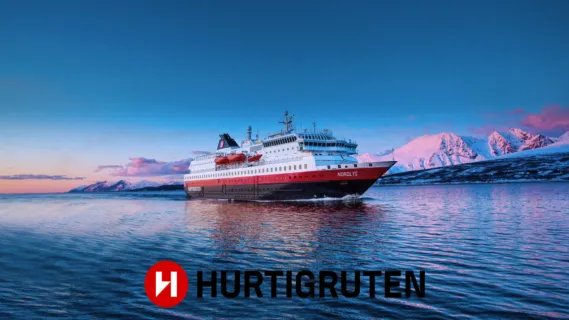 Hurtigruten Das Original: Authentische Postschiffreise entlang der norwegischen Fjordküste bei Sonnenuntergang. Ein klassisches Postschiff von Hurtigruten Das Original navigiert durch ruhige, tiefblaue Gewässer vor einer Kulisse aus schneebedeckten norwegischen Bergen im rosa Abendlicht der Dämmerung.