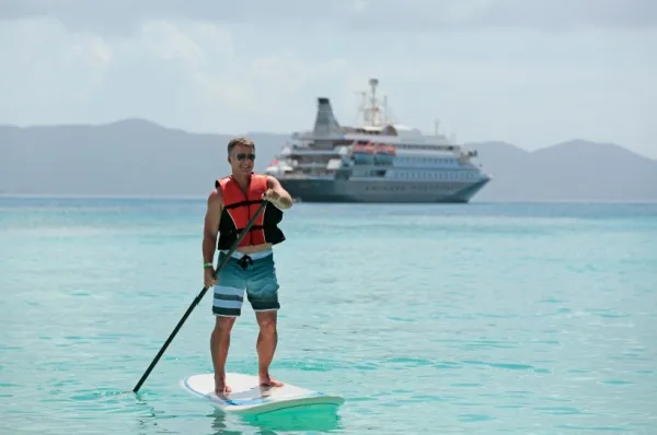 Sport bei SeaDream Yacht Club Ein Mann mit Schwimmweste auf einem Stand up Paddle in klarem Wasser. Im Hintergrund schwimmt die SeaDream I von SeaDream Yacht Club.