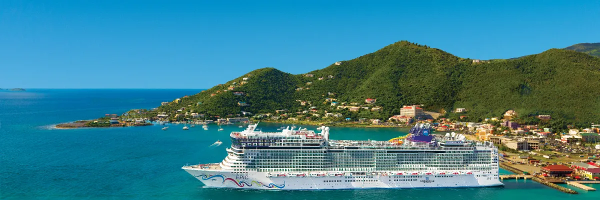 Luftaufnahme von Norwegian Epic, die im kristallklaren, türkisfarbenen Wasser vor einer üppig grünen Karibikinsel mit bunten Häusern vor Anker liegt.