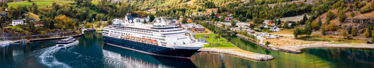 Panorama-Aufnahme des Kreuzfahrtschiffes VASCO DA GAMA von nicko cruises im Hafen eines norwegischen Fjords, flankiert von grünen Berghängen und einem kleinen Ausflugsboot auf dem glitzernden Wasser.
