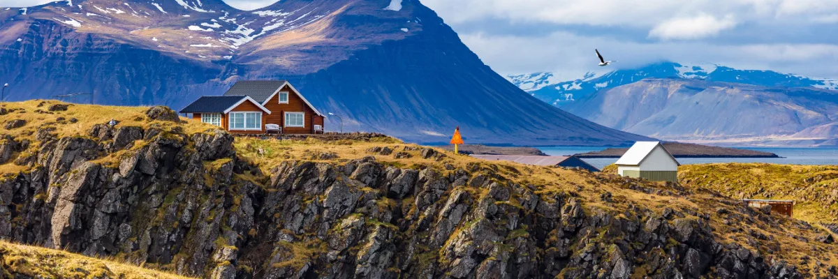 Ein einsames skandinavisches Holzhaus auf einer schroffen Felsklippe vor einer Kulisse aus gewaltigen, schneebedeckten Bergen und dem tiefblauen Nordmeer – typische Küstenlandschaft Islands.