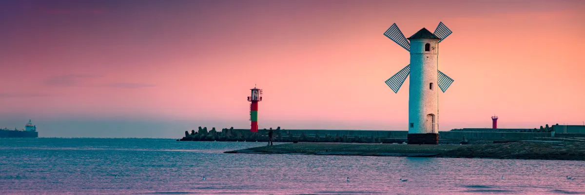 Die markante weiße Mühlenbake von Swinemünde auf einer Mole bei Sonnenuntergang, im Hintergrund ein rot-grüner Leuchtturm unter einem violett-rosa Abendhimmel an der Ostseeküste.