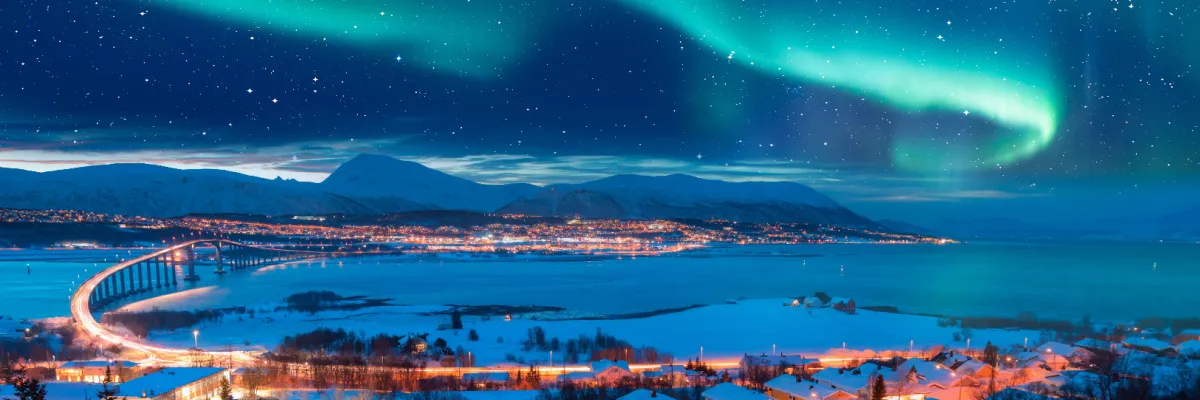 Spektakuläre grüne Nordlichter (Aurora Borealis) am sternenklaren Nachthimmel über einer hell beleuchteten Küstenstadt und einer markanten Brücke in Nordnorwegen während einer Winterkreuzfahrt.