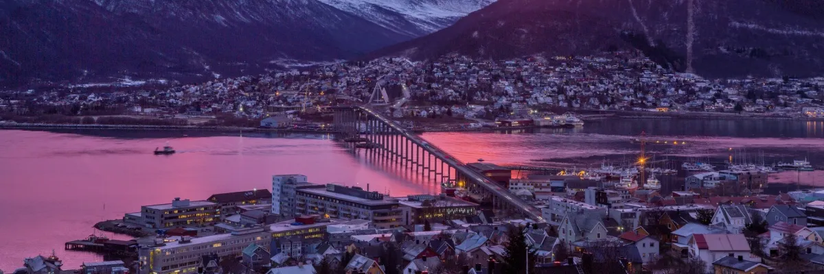 Die Stadt Tromsø in einem pink-violetten Sonnenuntergang. Eine Brücke führtt über einen Fjord.