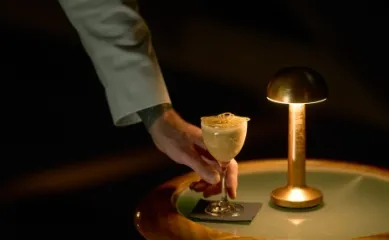 Eine Person vom Servicepersonal bei Hurtigruten stellt eine Kaffeekreation auf einen Beistelltisch neben eine goldene Lampe. ©Hurtigruten, Max Emanuelson