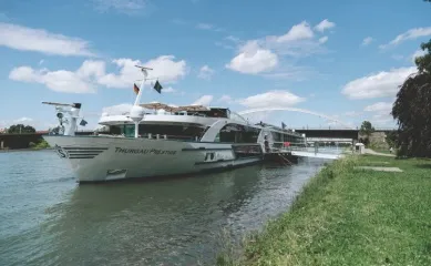 Die MS Thurgau Prestige macht Halt an einem Anleger am Ufer vor Wiesenlandschaften unter blauem Himmel.
