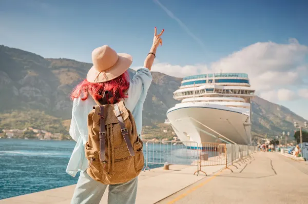 Eine Reisende mit Rucksack und Hut steht am Kai und blickt bei strahlendem Sonnenschein auf ein großes weißes Kreuzfahrtschiff im Hafen – Symbol für Vertrauen und Vorfreude bei Ahoi-Schiff.de.