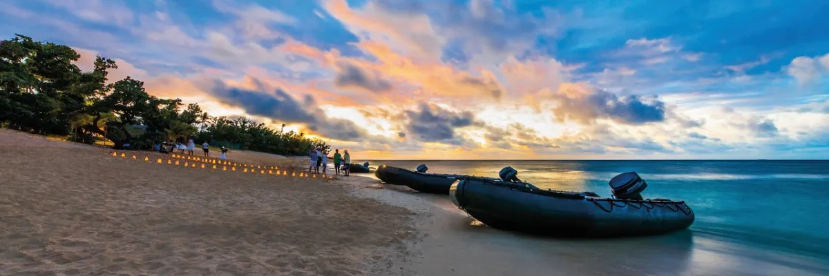 Ein stimmungsvoller Blick auf einen tropischen Strand während der Dämmerung. Mehrere schwarze Zodiac-Expeditionsboote von Hapag-Lloyd Cruises liegen am Ufer bereit, während Gäste den Abend unter einem dramatischen, orange-blau leuchtenden Himmel genießen.