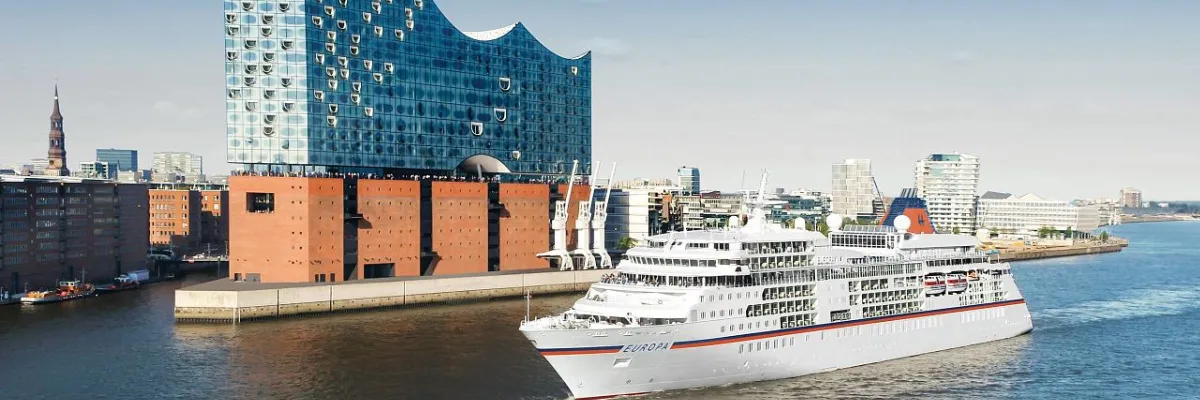 Das luxuriöse Kreuzfahrtschiff EUROPA von Hapag-Lloyd Cruises passiert die gläserne Fassade der Elbphilharmonie im Hamburger Hafen. Eine strahlend blaue Himmelsszenerie betont die Eleganz des Schiffes bei der Fahrt auf der Elbe.