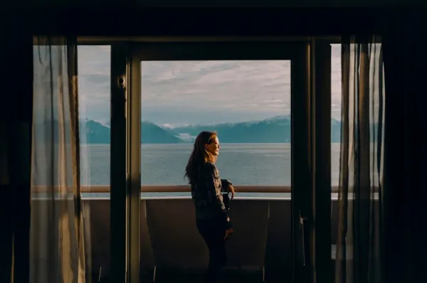 Eine Frau steht auf dem Balkon ihrer Expedition Suite auf MS Fridtjof Nansen zwischen zwei Gardinen und schaut auf die vorbeiziehende Landschaft.