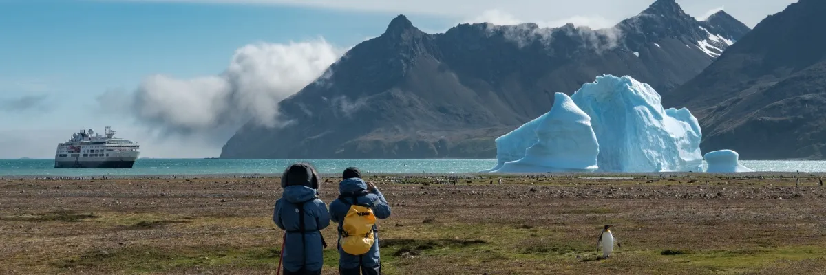 Zwei Personen stehen auf einer trockenen Wiese und schauen auf einen schwimmenden Gletscher und Berge in Südgeorgien, Fortuna Bay. Die MS Fram schwimmt im Hintergrund.