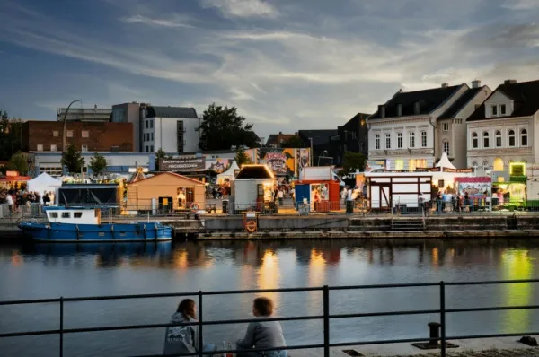 Ein Pärchen sitzt am Ufer der Elbe und schaut auf das geschäftige Treiben auf der anderen Uferseite, auf dem Büdchen mit Licht-Schilder aufgebaut sind.