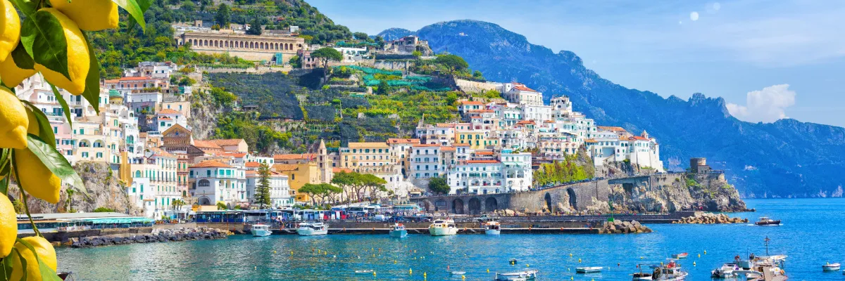 Mittelmeer-Highlights 2028: Mediterrane Schätze und kulinarische Träume entdecken Gelbe Zitronen hängen an einem Zweig im Vordergrund vor der malerischen Kulisse einer bunten Küstenstadt am Mittelmeer, die sich an die Klippen schmiegt. Das tiefblaue Wasser und der strahlende Himmel symbolisieren das „La Dolce Vita“.