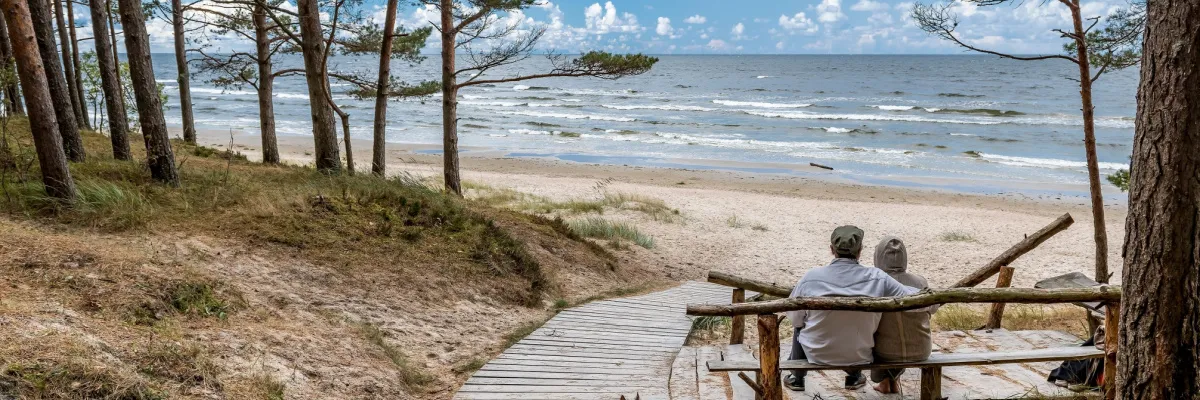 Maritime Auszeit 2028: Die schönsten Küsten der Ostsee entdecken Paar auf einer Holzbank blickt durch Bäume auf den Ostseestrand; Sinnbild für Erholung und Landgänge in Skandinavien oder Polen.