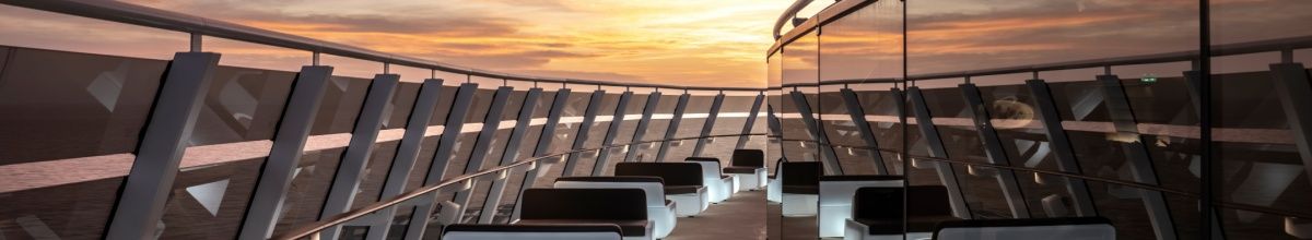 Blick auf die Top Sail Lounge, einen exklusiven Bereich für Gäste des MSC Yacht Club auf der MSC World Europa. Im Vordergrund der Außenbereich mit Sesseln und Tischen sowie Panoramascheiben. Im Hintergrund Sonnenuntergang über dem Meer.