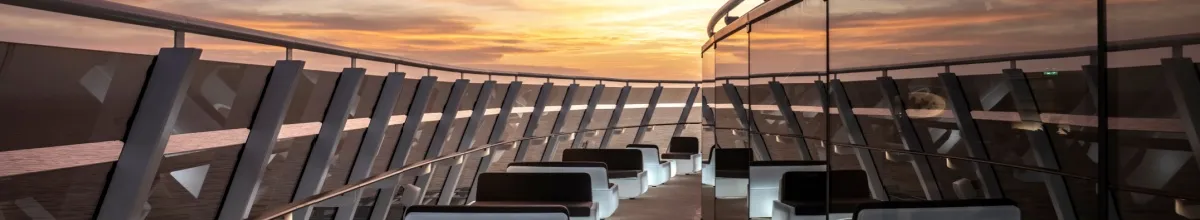 Gestalten Sie Ihre Reise mit den MSC Erlebniswelten Blick auf die Top Sail Lounge, einen exklusiven Bereich für Gäste des MSC Yacht Club auf der MSC World Europa. Im Vordergrund der Außenbereich mit Sesseln und Tischen sowie Panoramascheiben. Im Hintergrund Sonnenuntergang über dem Meer.