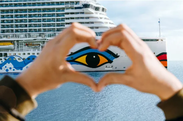 Hände formen ein Herz und rahmen das ikonische AIDA Auge am Bug eines Kreuzfahrtschiffes ein.