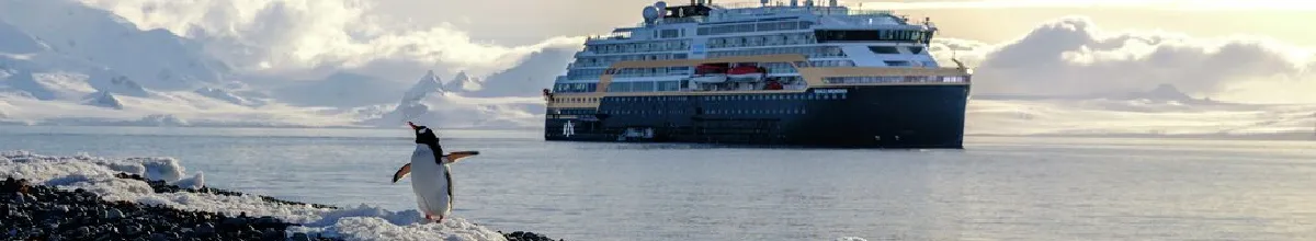 Ein Pinguin am verschneiten Ufer beobachtet ein modernes HX Expeditionsschiff in der eisigen Polarlandschaft der Antarktis zum 130-jährigen Jubiläum.