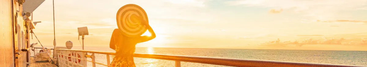 Entspannter Kreuzfahrt-Urlaub auf hoher See Eine Frau mit einem großen Sommerhut blickt von der Reling eines Kreuzfahrtschiffes auf den weiten Ozean im goldenen Abendlicht.