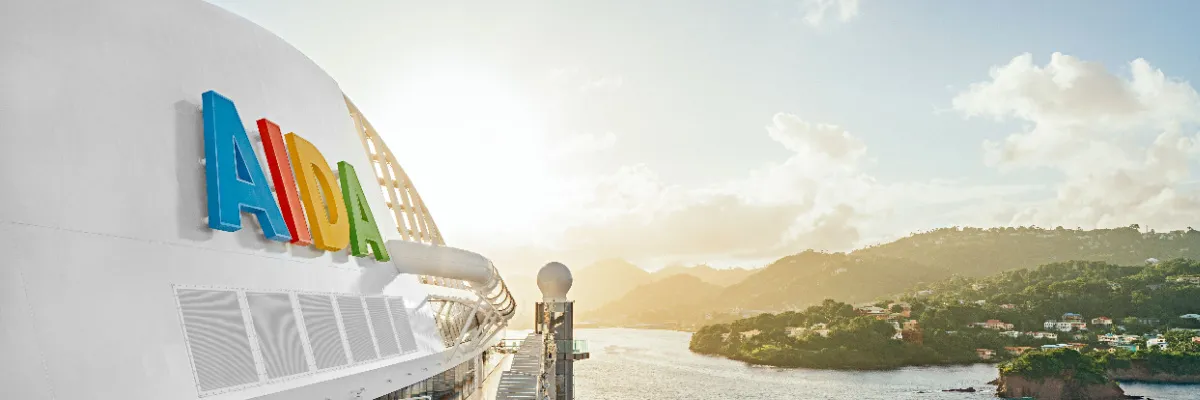 Nahaufnahme des bunten AIDA Logos an Bord eines Kreuzfahrtschiffes mit Blick auf eine sonnenbeschienene, grüne Inselbucht.