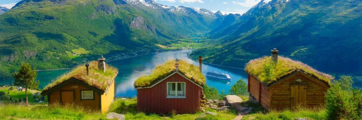 Atemberaubende Panorama-Aussicht von oben auf einen tiefblauen norwegischen Fjord, durch den ein Kreuzfahrtschiff gleitet. Im Vordergrund stehen drei urige Holz-Hütten mit typisch bewachsenen Grasdächern vor einer majestätischen Gebirgskulisse.