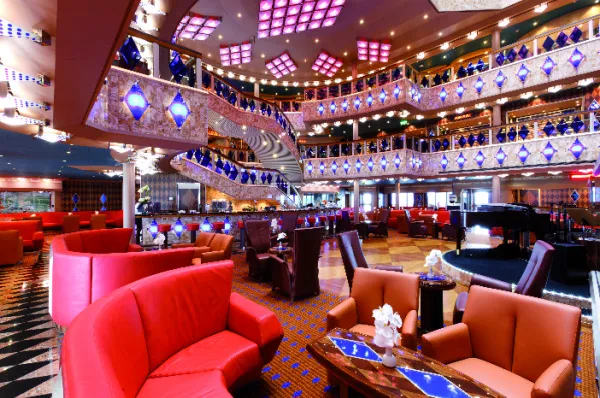 Das imposante, mehrstöckige Atrium der Costa Favolosa mit elegant geschwungenen Treppen, rot-orangefarbenen Polstersesseln, dekorativen blauen Leuchtelementen an den Balustraden und einer kleinen Showbühne samt Flügel.