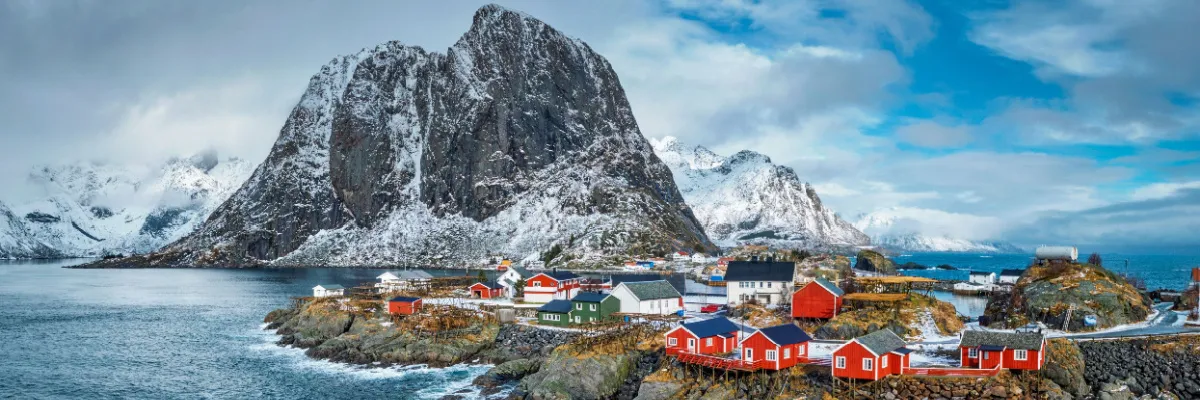 Traditionelle rote Fischerhütten an der felsigen Küste Norwegens vor einer Kulisse aus majestätischen, schneebedeckten Bergen.