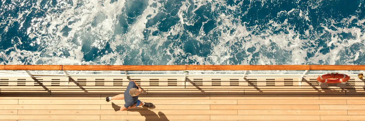 Aktive Erholung auf dem Promenadendeck – Sport und Fitness an Bord von Cunard Ein sportlicher Gast joggt auf dem edlen Teakdeck eines Cunard-Luxusschiffs, aufgenommen aus der Vogelperspektive direkt an der Reling neben tiefblauem Meereswasser.