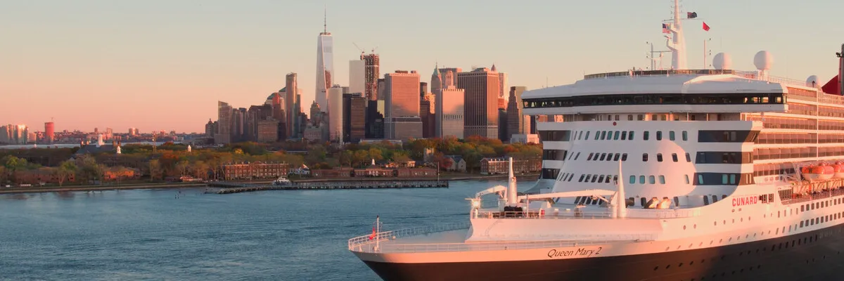 Die Queen Mary 2 in New York – Cunard Frühbucherangebote 2028/2029 Das legendäre Cunard-Flaggschiff die Queen Mary 2 läuft im goldenen Abendlicht vor der weltberühmten Skyline von New York City und dem One World Trade Center ein.