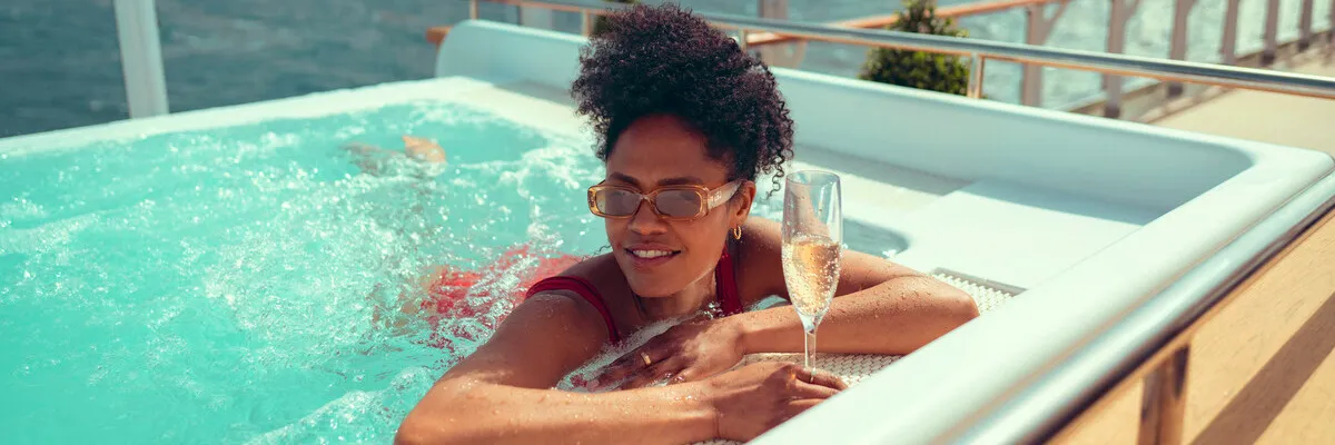 Exklusiver Komfort und Entspannung an Bord – Cunard Frühbucherangebote 2028/2029 Eine Dame mit Sonnenbrille entspannt sich mit einem Glas Champagner im Whirlpool auf dem weitläufigen Außendeck eines Cunard-Luxusschiffs.