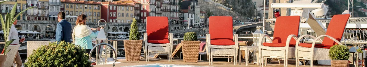 Das weitläufige Sonnendeck eines modernen A-ROSA Flusskreuzfahrtschiffes mit bequemen roten Liegestühlen vor einer malerischen historischen Flusskulisse.