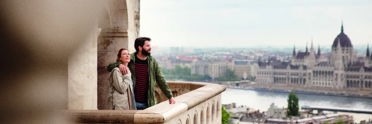 Ein lächelndes Paar steht auf einer historischen Steinterrasse in Budapest und blickt über die herbstliche Donau auf das majestätische Parlamentsgebäude.
