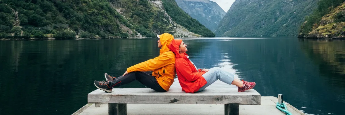 Ein Paar in wetterfesten, farbenfrohen Outdoorjacken sitzt Rücken an Rücken auf einem Holzsteg vor einem stillen norwegischen Fjord.