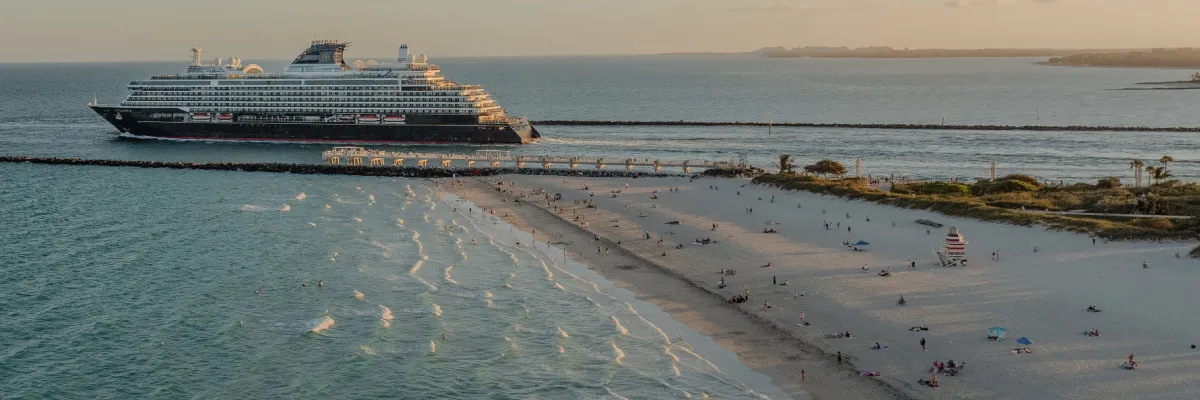 Das elegante Luxusschiff von Explora Journeys gleitet im warmen Abendlicht an einem feinen Sandstrand mit Palmen und Badegästen vorbei aufs offene Meer.