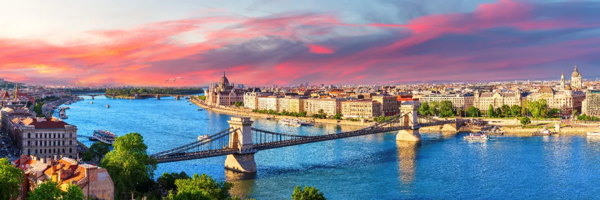 Budapest an der Donau mit Kettenbrücke – nicko cruises Donaukreuzfahrten Panorama von Budapest an der Donau mit der berühmten Kettenbrücke und dem Parlamentsgebäude unter einem farbenfrohen Abendhimmel beim nicko cruises Sommerflussverkauf.