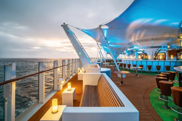 Gemütliche Abendstunden an Deck von AIDAnova während der Eventreise Crime & Sea Elegante Außenbar an Deck von AIDAnova mit blau beleuchtetem Segeldach, einladenden Loungemöbeln und Blick auf den herbstlich-winterlichen Ozean bei Sonnenuntergang.