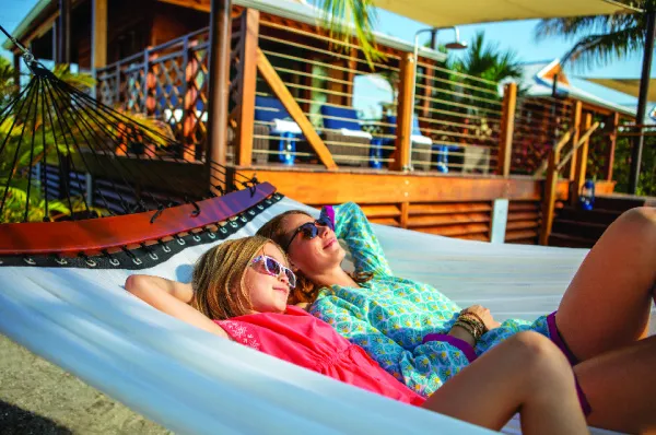 Harvest Caye Strandvillen – Komfortabler Landausflug in Belize Eine Frau und ein Mädchen entspannen in einer weißen Hängematte vor einer exklusiven hölzernen Strand-Cabana mit Terrasse auf Harvest Caye.