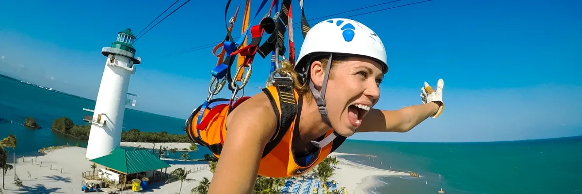 Nervenkitzel auf Harvest Caye – Zipline-Flug am Flighthouse-Tower Eine begeisterte Urlauberin im Flug an der Zipline auf Harvest Caye vor der Kulisse des markanten weißen Leuchtturms und des türkisblauen Meers.