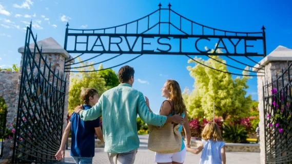 Eine Familie läuft unter dem großen, schwarzen Eingangstor mit dem geschwungenen Schriftzug „Harvest Caye“ auf der tropischen Privatinsel in Belize.