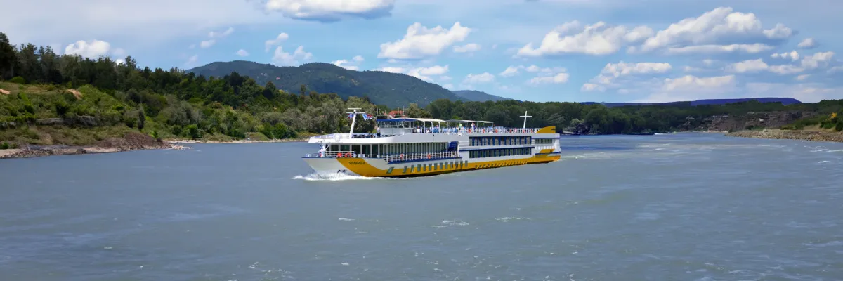 Ein komfortables 1AVista-Flussschiff auf Kurs durch idyllische Landschaften Ein modernes Flusskreuzfahrtschiff von 1AVista Reisen im markanten gelb-weißen Design navigiert auf einem breiten Fluss vor einer grünen, waldreichen Hügelkette unter blauem Himmel.