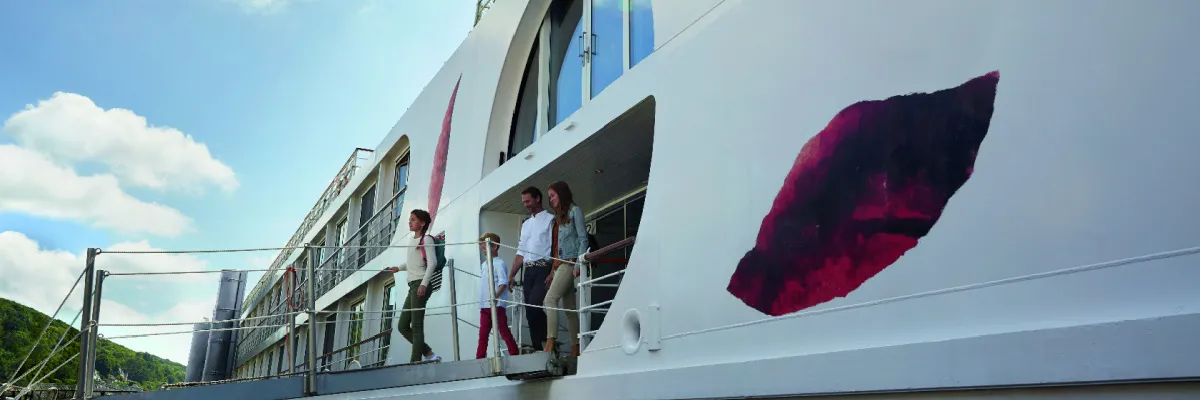 Sorgloser Familienurlaub und Landgang auf einer A-ROSA Flusskreuzfahrt Gäste gehen über die Gangway von Bord eines eleganten, weißen A-ROSA Flusskreuzfahrtschiffes, auf dessen Außenwand das ikonische rote Kussmund-Logo der Reederei zu sehen ist.