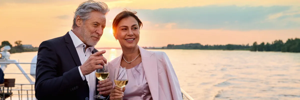 Stilvoller Genuss bei Sonnenuntergang an Bord von AMADEUS River Cruises Ein elegant gekleidetes Paar steht am Abend auf dem Außendeck eines Premium-Flusskreuzfahrtschiffes, hält Gläser mit Weißwein und genießt den weiten Blick über den Fluss im goldenen Sonnenuntergang.