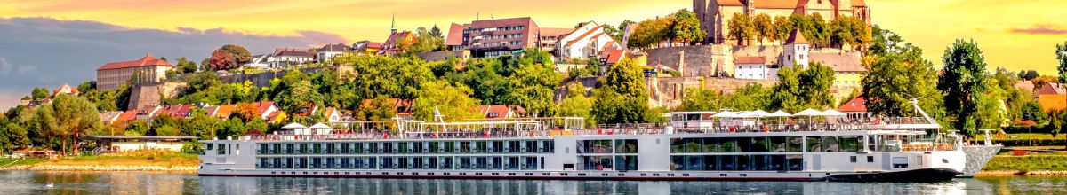 Ein elegantes, weißes Flusskreuzfahrtschiff der Premium-Klasse fährt bei goldenem Abendlicht auf dem Fluss vor einer malerischen Kulisse mit historischer Burg und Altstadt auf einem grünen Hügel.