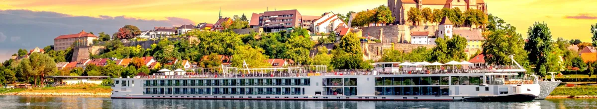 Ein elegantes, weißes Flusskreuzfahrtschiff der Premium-Klasse fährt bei goldenem Abendlicht auf dem Fluss vor einer malerischen Kulisse mit historischer Burg und Altstadt auf einem grünen Hügel.