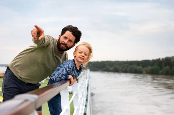 Gemeinsame Familienmomente an Deck während einer Flusskreuzfahrt Ein Vater und sein Sohn stehen an der Reling eines Flusskreuzfahrtschiffes und blicken lächelnd auf den Fluss, während der Mann in die Ferne zeigt.
