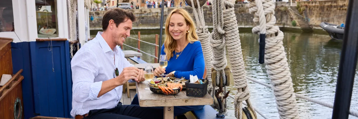Exklusiver kulinarischer Genuss und Premium-Lifestyle an Bord von VIVA Cruises Ein lächelndes Paar sitzt an einem rustikalen Holztisch auf dem Außendeck eines Schiffes, genießt eine frische Meeresfrüchteplatte und stößt mit Gläsern Weißwein an.